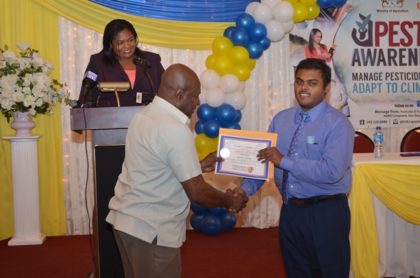 Chairman of the PTCCB, Dr. Leslie Munroe presents a certificate to a graduate, at the PTCCB Graduation Ceremony as Registrar of the PTCCB, Trecia Garnath (in Background) looks on 